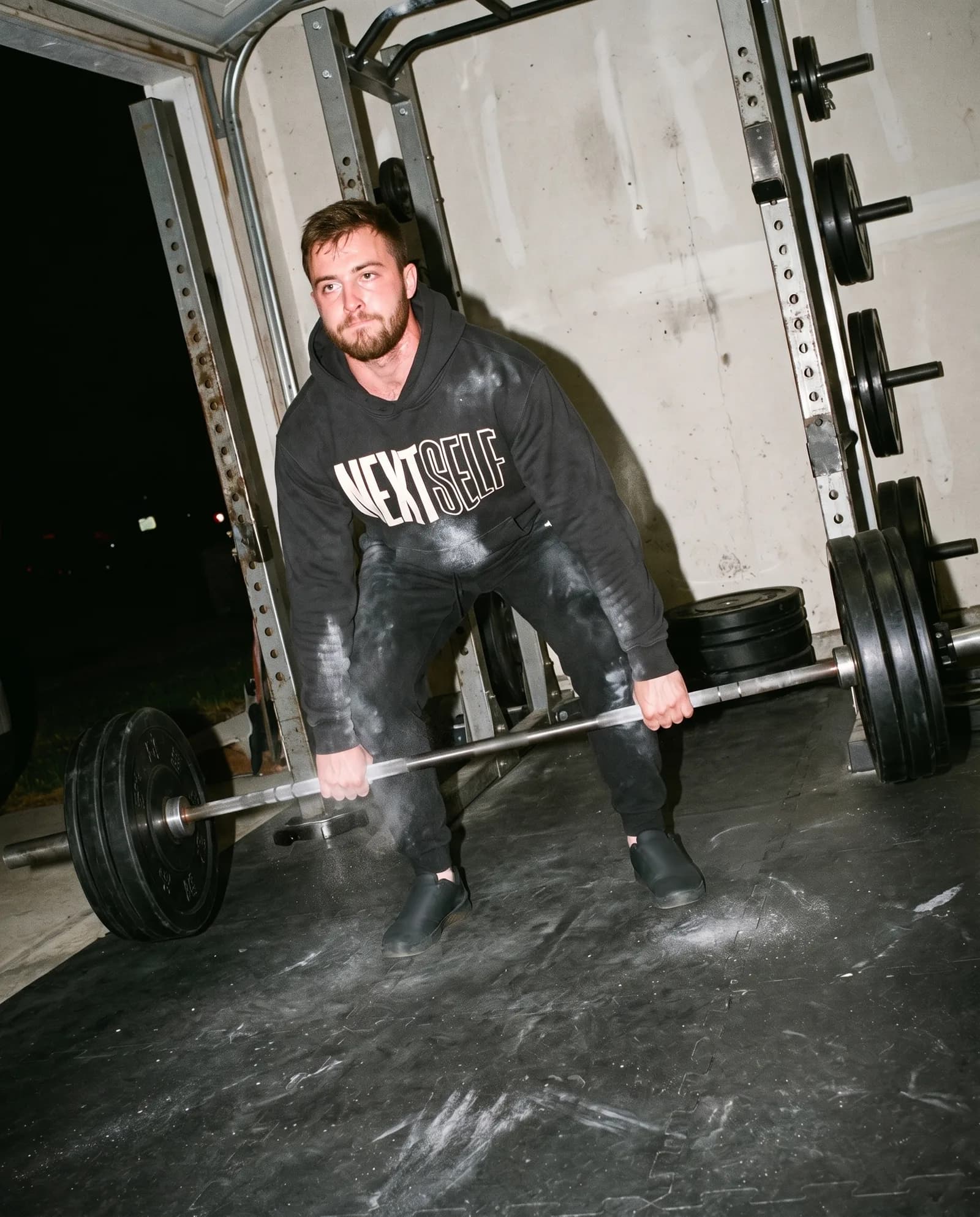 A lifter at deadlift lockout in a garage gym at night, wearing the black Outline Hoodie